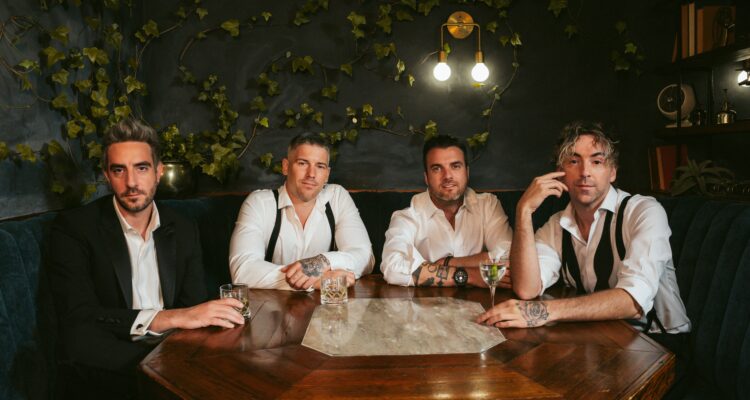 Promotional-style photo of four All Time Low band members seated around a wooden table in a dim, intimate lounge with ivy on the dark walls. They wear white shirts, some with suspenders or jackets, and hold drinks as they pose under warm hanging lights.